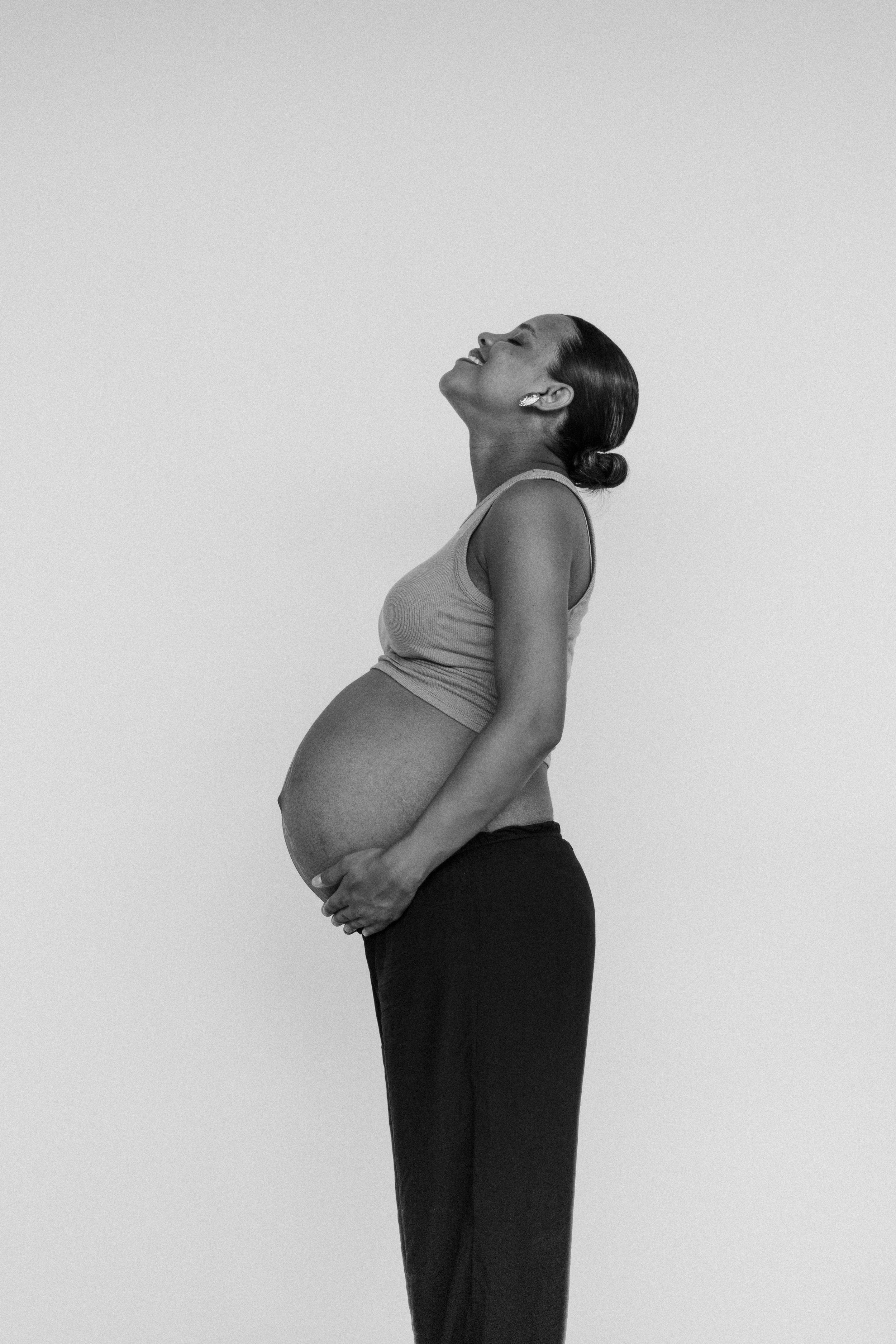 A black-and-white photo of a pregnant woman smiling with her head tilted back, wearing earrings and a tank top, standing against a plain background.